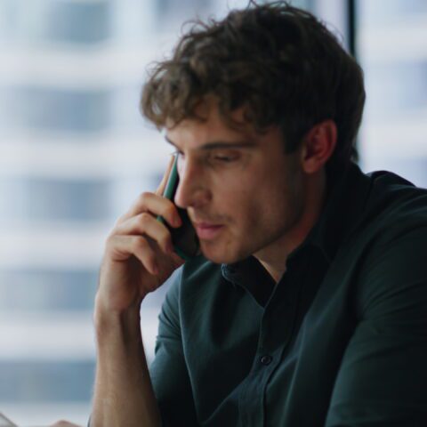 John DOE 1 Homme concentré passant un appel téléphonique à son bureau devant une baie vitrée.