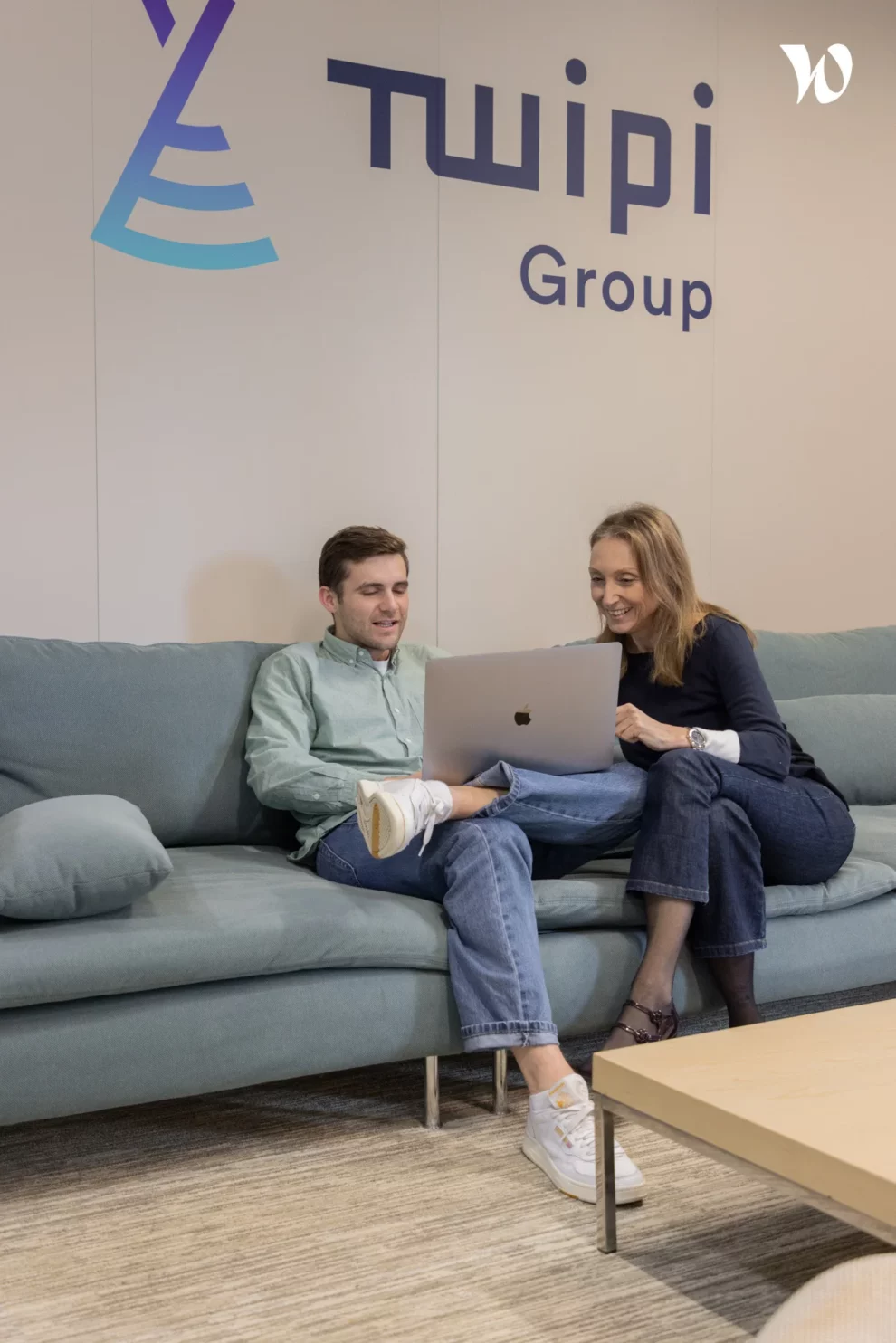 Deux collaborateurs de Twipi Group échangeant devant un ordinateur portable sous le logo mural de l'entreprise.