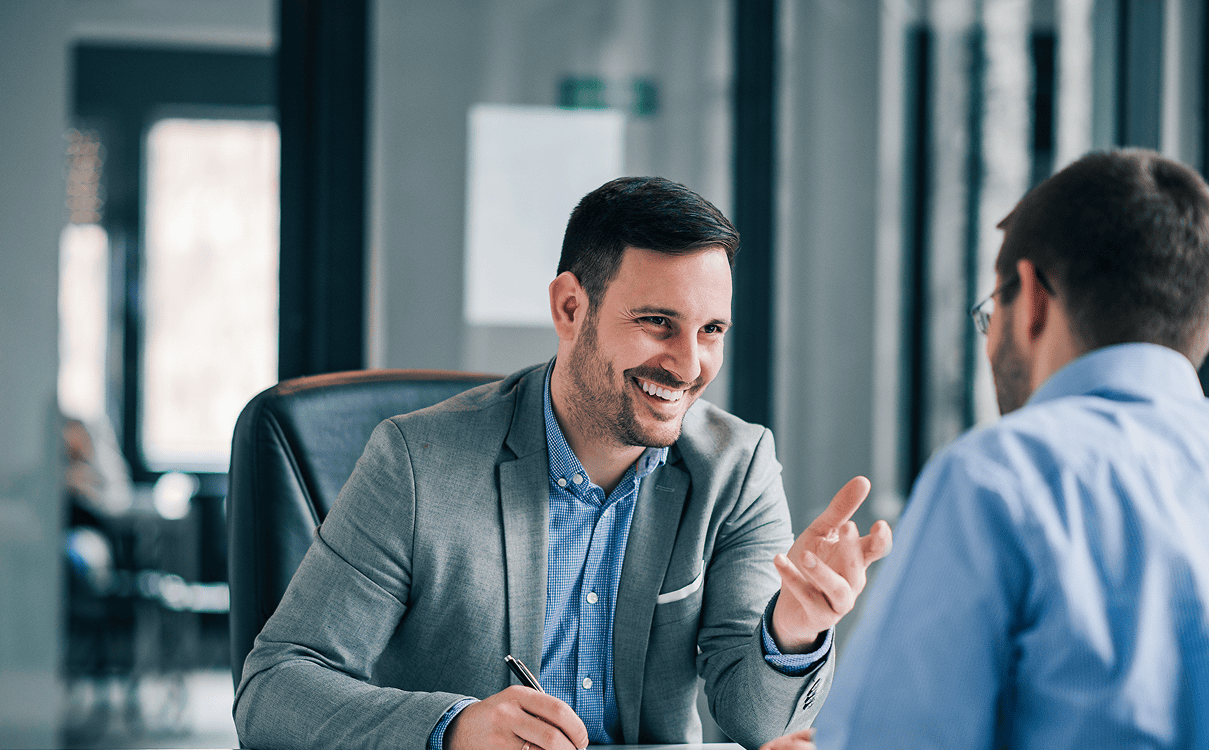 Homme souriant en costume en pleine discussion avec un interlocuteur à son bureau.