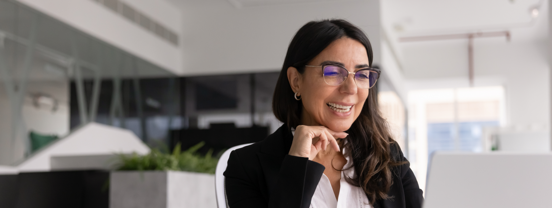 Femme souriante en tailleur, main au menton, à son bureau dans un espace de travail.