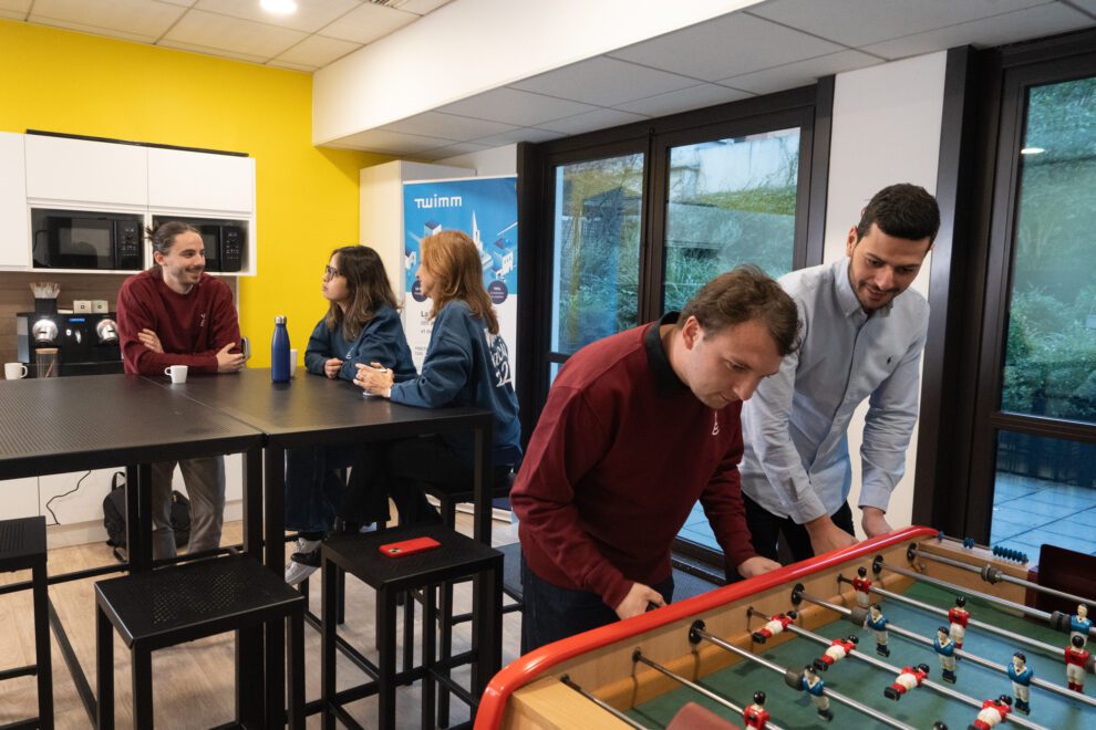 Nous rejoindre 1 Collaborateurs de Twipi Group jouant au baby-foot dans l'espace détente, devant un kakémono Twimm.