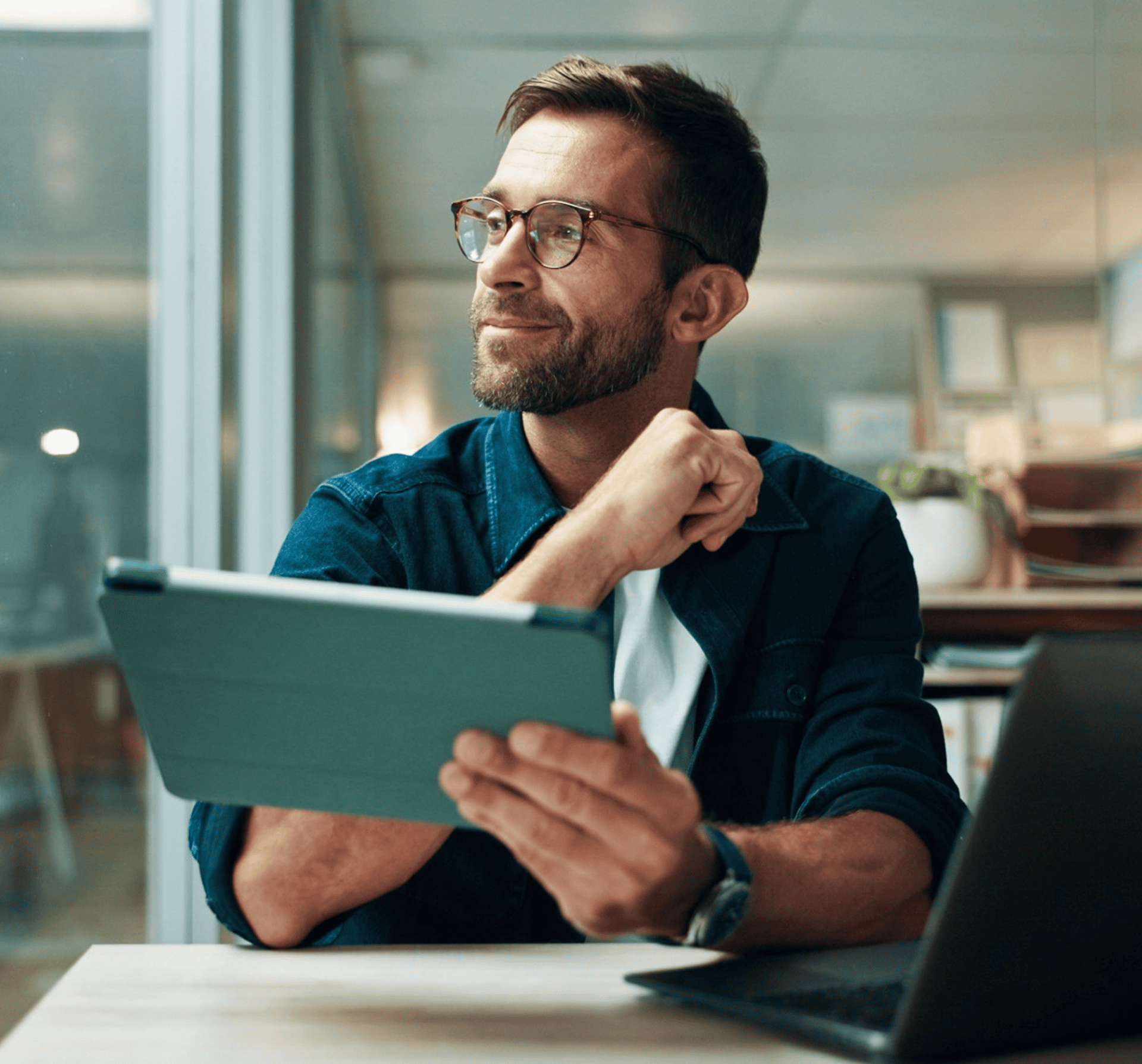 Homme à lunettes assis à son bureau avec une tablette, dans un espace de travail.